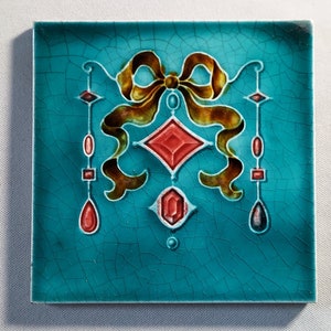 Może przedstawiać: Kwadratowa płytka ceramiczna z turkusowym tłem i dekoracyjnym wzorem. Wzór przedstawia brązową i złotą kokardę, czerwone kształty rombów i akcenty w kształcie łezki. Płytka ma spękaną fakturę.