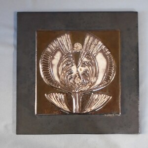 May include: A square ceramic wall tile featuring a raised, glossy brown and cream relief of a stylized flower. The central floral design is set against a dark, matte-finished square border, creating a distinct contrast between the textures.