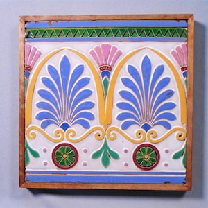 May include: Square decorative tile with a wooden frame. The tile features a symmetrical design with blue, yellow, pink, green, and white elements. The central motif includes stylized blue leaf-like shapes within yellow arches.