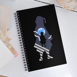 May include: Black spiral-bound notebook featuring a silhouette design with a girl, a moon, and snakes. The design is in shades of blue and white against a black background. The notebook is on a white surface.