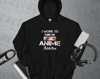 Puede incluir: Sudadera con capucha negra con el texto blanco "I Work To Fund My Anime Addiction". El diseño incluye ojos de anime. Un par de zapatillas negras y un smartphone con auriculares también están en la imagen.