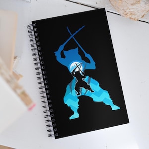 May include: Black spiral-bound notebook featuring a blue and black graphic design. The design depicts a silhouette of a figure holding swords, with a moon and mountain-like shapes in the background. The notebook has a white spiral binding.