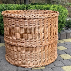 May include: A large, cylindrical, light brown woven wicker basket featuring a sturdy braided rim at the top and a matching decorative braided band encircling the middle section. The basket is constructed from natural reed with a tight weave.