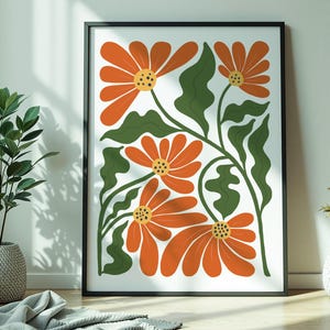 May include: A framed floral art print featuring orange flowers with yellow centers and green leaves against a white background. The artwork is in a black frame and leans against a white wall. The room also has plants and vases.
