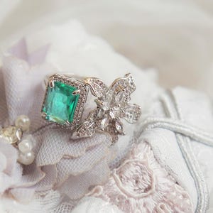 Op de afbeelding: Een ring met een smaragdgroene edelsteen met smaragd-slijpsel en bloemendetails, geplaatst op witte en lavendelkleurige stof met parels en kant. De ring is het middelpunt en toont zijn ingewikkelde details en sprankelende accenten.