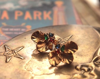 Puede incluir: Un espejo de mano dorado de estilo vintage con diseño de media luna. Dos pendientes florales dorados con gemas de colores descansan sobre el espejo. Una postal vintage con las palabras "LUNA PARK" está en el fondo.