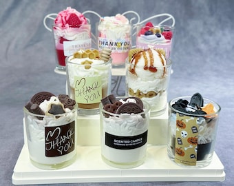 Puede incluir: Una colección de ocho velas pequeñas en vaso dispuestas sobre un soporte blanco de varios niveles. Cada vela presenta adornos de cera que parecen postres, con etiquetas que dicen Thank You o Scented Candle.