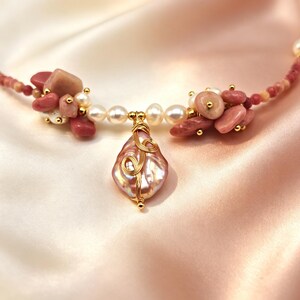 Op de afbeelding: Een ketting met een centrale druppelvormige iriserende parel omwikkeld met gouddraad, geflankeerd door clusters van roze steentjes en witte parels. Het snoer bestaat uit kleine roze kralen met overal goudkleurige accenten in het ontwerp.