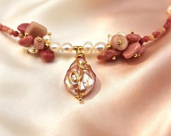 Puede incluir: Un collar con una perla central iridiscente en forma de lágrima envuelta en alambre dorado, flanqueada por grupos de piedras rosas y perlas blancas. El hilo consta de pequeñas cuentas rosas con detalles en tono dorado en el diseño.