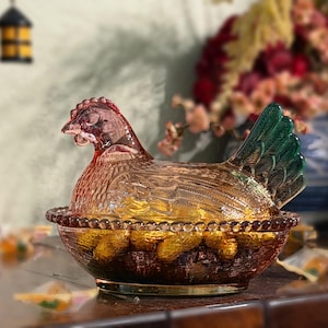 Op de afbeelding: Een glazen schaal in de vorm van een kip op een nest van geperst glas met een doorschijnend amberkleurig lichaam en een opvallende groene staart. Het deksel toont een kip met gevederde textuur op een kom met een rand van glazen kralen.