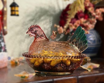 Puede incluir: Un plato de vidrio prensado con forma de gallina sobre un nido, con un cuerpo de color ámbar translúcido y una cola verde distintiva. La tapa tiene forma de gallina con detalles de plumas texturizadas sobre una base redonda con borde perlado.