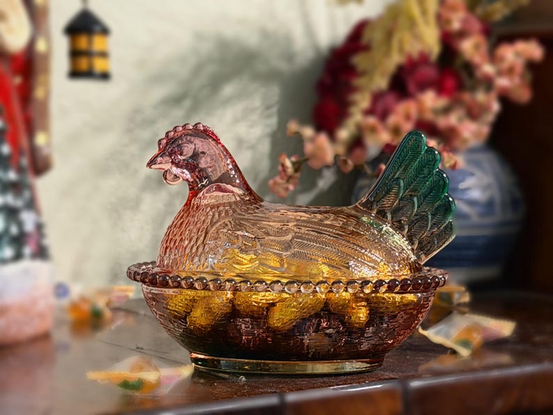 Op de afbeelding: Een glazen schaal in de vorm van een kip op een nest van geperst glas met een doorschijnend amberkleurig lichaam en een opvallende groene staart. Het deksel toont een kip met gevederde textuur op een kom met een rand van glazen kralen.
