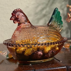 Op de afbeelding: Een glazen schaal in de vorm van een kip op een nest, gemaakt van doorschijnend amberkleurig glas met een gestructureerd verenpatroon en een felgroene staart. De basis van de schaal heeft een kralenrand en bevat goudkleurige snoepjes.