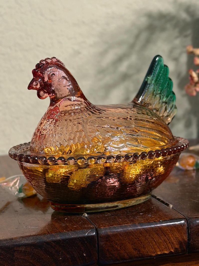 Op de afbeelding: Een glazen schaal in de vorm van een kip op een nest, gemaakt van doorschijnend amberkleurig glas met een gestructureerd verenpatroon en een felgroene staart. De basis van de schaal heeft een kralenrand en bevat goudkleurige snoepjes.