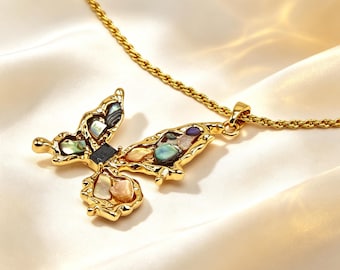 Puede incluir: Un collar de tono dorado con un colgante en forma de mariposa. El colgante tiene un marco de metal dorado texturizado relleno con incrustaciones de fragmentos de concha iridiscentes en tonos azul, verde, rosa y blanco, centrado por un pequeño cuadrado negro.