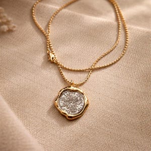 Op de afbeelding: Een gouden ketting met een ronde hanger met een zilverkleurige munt in het midden. De hanger heeft een onregelmatige, golvende gouden rand rondom de getextureerde zilveren munt, die voorzien is van reliëfdetails en letters.