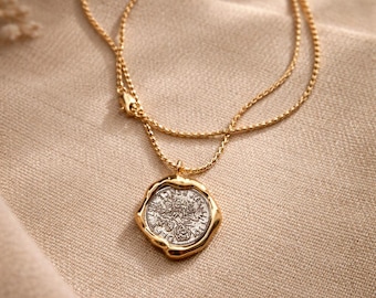 Puede incluir: Un collar de cadena dorada con un colgante redondo que tiene una moneda plateada en el centro. El colgante presenta un borde dorado irregular y ondulado que rodea la moneda de plata texturizada, con detalles en relieve y letras.
