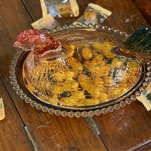 Op de afbeelding: Een amberkleurige glazen schaal in de vorm van een kip op een nest met een getextureerd lichaam, een rood getinte kop en een groen getinte staart. Het deksel heeft een gedetailleerde kippenvorm en de basis is voorzien van een parelrand.