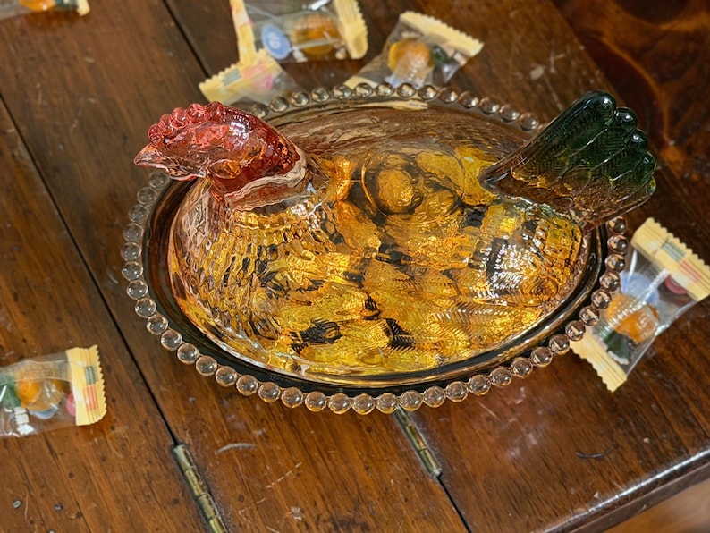 Op de afbeelding: Een amberkleurige glazen schaal in de vorm van een kip op een nest met een getextureerd lichaam, een rood getinte kop en een groen getinte staart. Het deksel heeft een gedetailleerde kippenvorm en de basis is voorzien van een parelrand.