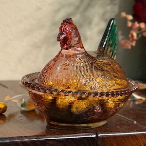 Op de afbeelding: Een glazen schaal in de vorm van een kip op een nest, gemaakt van geperst glas met een amberkleurig lichaam, een textuur van veren, een rand met kralenpatroon en een opvallende groene staart. Het deksel is gevormd als een kip met een kop.