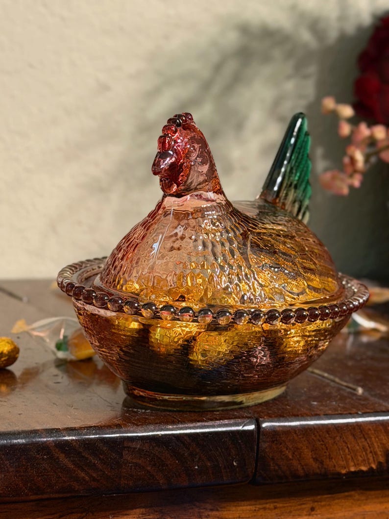 Op de afbeelding: Een glazen schaal in de vorm van een kip op een nest, gemaakt van geperst glas met een amberkleurig lichaam, een textuur van veren, een rand met kralenpatroon en een opvallende groene staart. Het deksel is gevormd als een kip met een kop.
