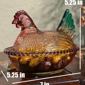 Op de afbeelding: Een doorschijnende amberkleurige glazen schaal in de vorm van een kip op een nest met een groene staart, voorzien van een kralenrand en gestructureerde veertjes. De schaal is 13,34 cm hoog en 17,78 cm lang, volgens de zichtbare maattekst.