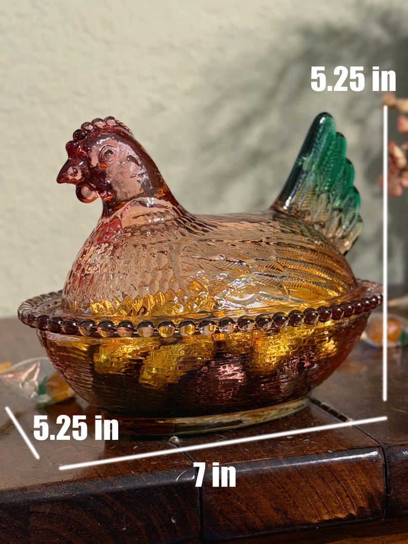 Op de afbeelding: Een doorschijnende amberkleurige glazen schaal in de vorm van een kip op een nest met een groene staart, voorzien van een kralenrand en gestructureerde veertjes. De schaal is 13,34 cm hoog en 17,78 cm lang, volgens de zichtbare maattekst.