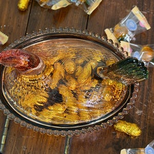 Op de afbeelding: Een amberkleurige glazen kip-op-nest schaal met een getextureerde, kralenrand. Het deksel is gevormd als een kip met een roodachtige kop en een donkergroene staart, voorzien van gedetailleerde verenpatronen. De ovale basis heeft een heldere textuur.