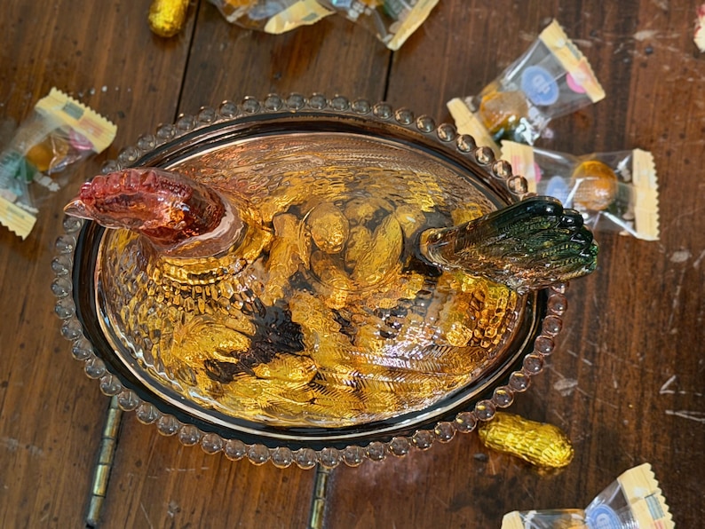Op de afbeelding: Een amberkleurige glazen kip-op-nest schaal met een getextureerde, kralenrand. Het deksel is gevormd als een kip met een roodachtige kop en een donkergroene staart, voorzien van gedetailleerde verenpatronen. De ovale basis heeft een heldere textuur.