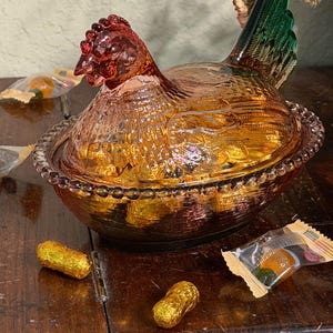 Op de afbeelding: Een glazen schaal in de vorm van een kip op een nest van persglas met een amberkleurig lichaam, een gestructureerd verenpatroon en een donkergroene staart. Het deksel is gevormd als een kip en de basis heeft een kralenrand. Goud verpakte snoepjes.
