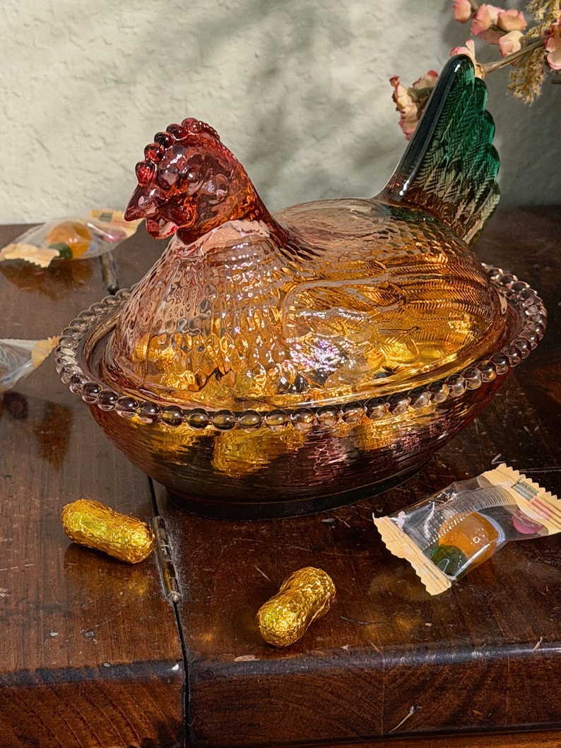 Op de afbeelding: Een glazen schaal in de vorm van een kip op een nest van persglas met een amberkleurig lichaam, een gestructureerd verenpatroon en een donkergroene staart. Het deksel is gevormd als een kip en de basis heeft een kralenrand. Goud verpakte snoepjes.