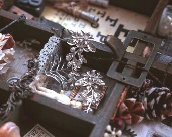 Puede incluir: Primer plano de una caja de madera llena de varios objetos de estilo vintage. Destacan accesorios para el cabello florales plateados con detalles de perlas. Otros elementos incluyen una hebilla de metal, encaje y una pequeña imagen enmarcada. La paleta de colores es apagada, con tonos marrones, plateados y blancos.