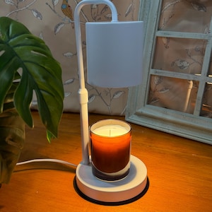 May include: A white electric candle warmer with a cylindrical shade and base. A lit candle in a glass jar sits on the base. The candle has a gradient orange color. The warmer is on a wooden surface.