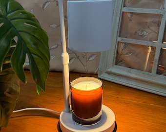 May include: A white electric candle warmer with a cylindrical shade and base. A lit candle in a glass jar sits on the base. The candle has a gradient orange color. The warmer is on a wooden surface.