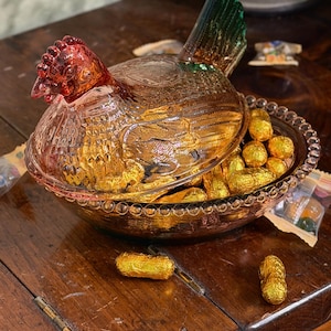 Op de afbeelding: Een geperste glazen schaal in de vorm van een kip op een nest met een doorschijnend amberkleurig lichaam, een donkerrode kop en een groene staart. Het deksel is gevormd als een kip en rust op een ronde kom met een rand van glazen kralen.