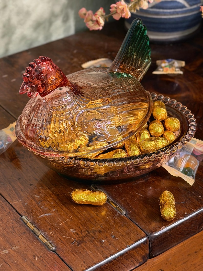 Op de afbeelding: Een geperste glazen schaal in de vorm van een kip op een nest met een doorschijnend amberkleurig lichaam, een donkerrode kop en een groene staart. Het deksel is gevormd als een kip en rust op een ronde kom met een rand van glazen kralen.