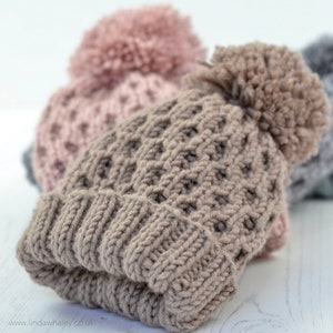May include: Three knitted hats with pom-poms in shades of pink, brown, and grey. The hats are made with a textured stitch and are stacked on top of each other. The text "Pure Hand Knits" is visible at the top of the image.