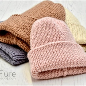 May include: Three knitted beanies in different colours: pink, brown, and cream. The beanies are folded and stacked on a white wooden surface. The text "Pure Hand Knits" is visible at the bottom of the image.