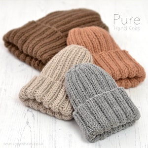 May include: Four hand-knitted beanies in different colours: brown, beige, peach, and grey. The beanies are stacked on top of each other on a white wooden surface. The text "Pure Hand Knits" is visible in the top right corner of the image.