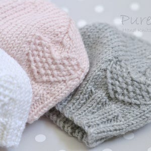 May include: Three hand-knitted baby hats in pink, white, and gray with a heart pattern. The hats are made with soft yarn and are perfect for newborns. Pure Hand Knits.