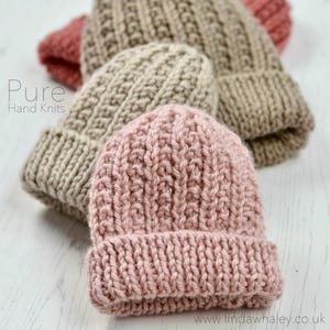 Puede incluir: Tres gorros de punto en diferentes colores: beige, marrón claro y rosa. Los gorros están hechos con un hilo grueso y tienen un diseño simple y clásico. El texto "Pure Hand Knits" es visible en la imagen.