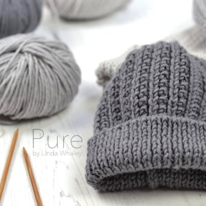 May include: A gray knitted beanie hat with a ribbed texture. The hat is made with a chunky yarn and has a folded brim. The text "Pure by Linda Whaley" is visible on the white surface below the hat.