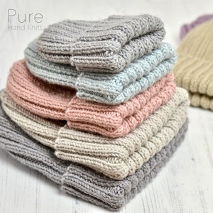 May include: A stack of five hand-knitted hats in various colors, including gray, light blue, pink, beige, and a hint of purple. The hats are folded and stacked on a white wooden surface. The text "Pure Hand Knits" is visible at the top of the image.