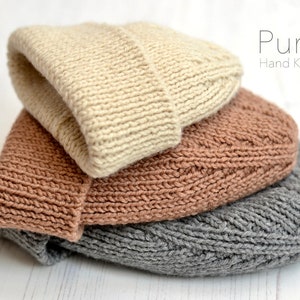 Könnte beinhalten: Drei gestrickte Mützen, die übereinander gestapelt sind. Die oberste Mütze ist beige, die mittlere Mütze ist braun und die unterste Mütze ist grau. Die Mützen sind alle handgestrickt und haben ein schlichtes, klassisches Design. Der Text "Pure Hand Knits" ist in der oberen rechten Ecke des Bildes sichtbar.