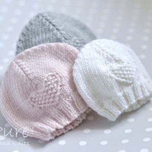 May include: Three knitted baby hats in pink, white, and gray. The hats are made with a textured stitch and have a simple design.