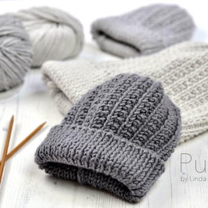 Peut inclure: Deux bonnets en tricot gris et deux pelotes de laine, une grise et une blanche, avec des aiguilles à tricoter sur une surface en bois blanc. Le texte "Pure by Linda Whaley" est visible dans le coin inférieur droit de l'image.