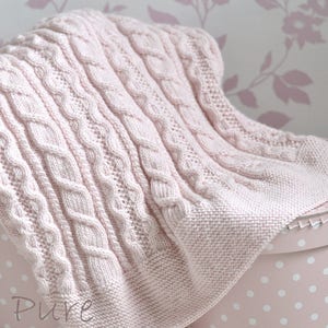 May include: A soft pink knitted baby blanket with a cable knit pattern. The blanket is draped over a pink and white polka dot box. The text "Pure Hand Knits" is visible in the lower left corner of the image.