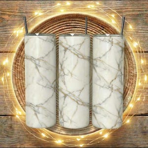 Może przedstawiać: Trzy białe kubki z marmurowym wzorem ze srebrnymi słomkami są prezentowane na tkanej macie, otoczone łańcuchem świetlnym. Kubki mają złoty wzór żyłkowy. Tło to drewniana powierzchnia.