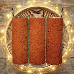 May include: Three cylindrical tumblers with a brick-like texture, each topped with a silver lid and a straw. The tumblers are arranged on a woven circular mat, with a string of warm white fairy lights around the mat. The background is a wooden surface.