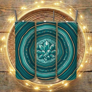 May include: Three teal-colored tumblers with a gemstone design. The design features concentric circles in shades of teal and gold, with a central crystal formation. Each tumbler has a silver straw. The tumblers are displayed on a woven mat.